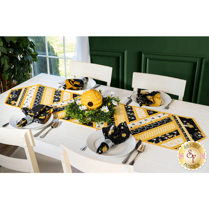 Table runner made of diagonal strips of bee themed fabrics in yellow, black, and white.