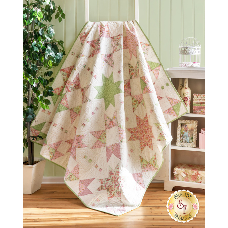 A quilt with geometric square and triangle designs made of pastel color fabrics on white draped over furniture.