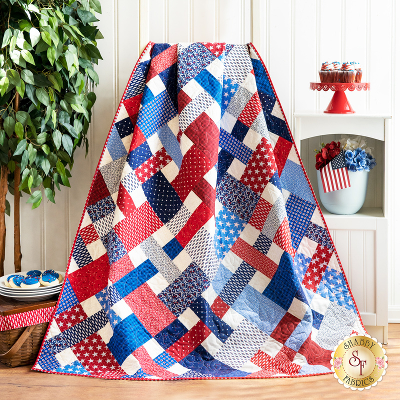 Red, white, and blue quilt with criss cross design made of patriotic printed fabrics draped over furniture.