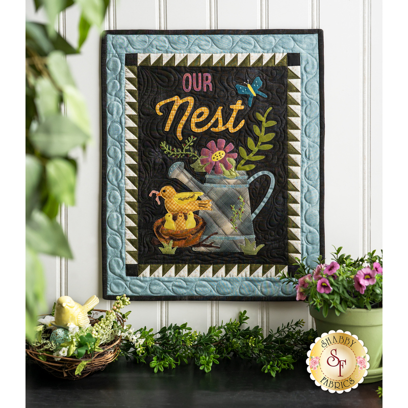 Wall hanging featuring appliqué of a nest of birds next to a watering can full of flowers and the phrase 