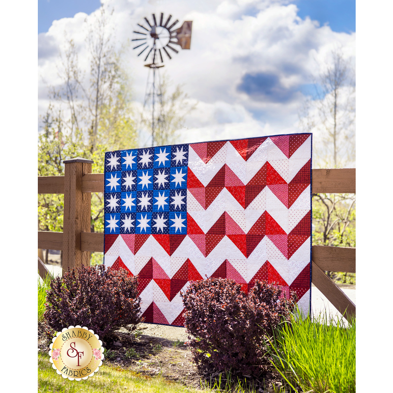 Quilted United States flag with chevron stripes and geometric stars outside on wood fence.