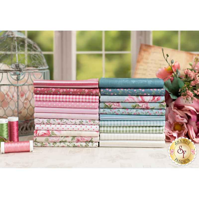 Stacks of pink, blue, and cream fabric on a white table with rusty pink roses, spools of thread, and other decorations in the background.