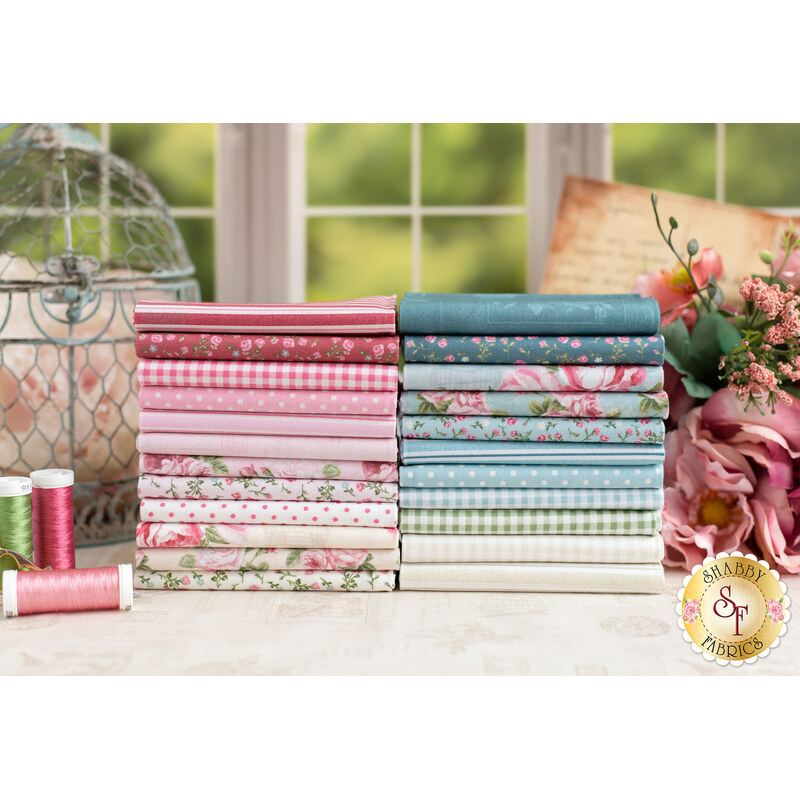 Stacks of pink, blue, and cream fabric on a white table with rusty pink roses, spools of thread, and other decorations in the background