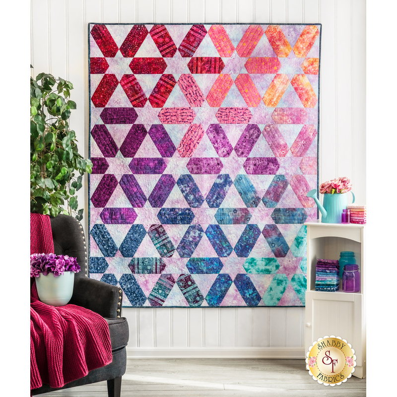 A colorful quilt with a geometric design hangs on a wall next to a cozy chair and shelf with fabric.