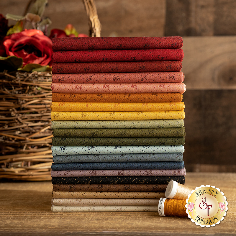A stack of red, brown, yellow, green, blue, and cream colored fabrics next to two spools of thread and a wicker basket