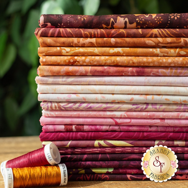 Close up photo of a stack of batik fabric arranged in a gradient of brown and pink with two spools of thread on the counter and greenery in the background.