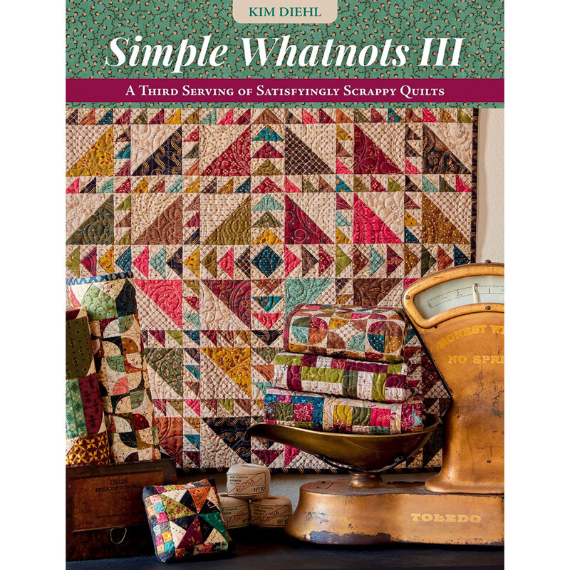 front cover of Simple Whatnots III featuring a finished quilt hanging on a wall in the background with an antique scale holding folded quilts on a tabletop in the foreground