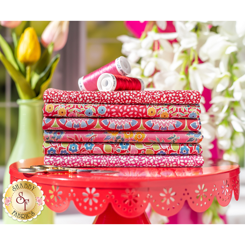 Stack of all of the fabrics in the Pie in the Sky - Red FQ Set, sitting on a small red table with spools of thread on top of them and flowers in the background