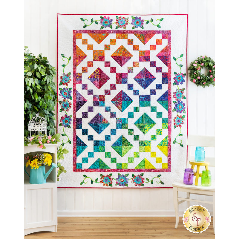A large white quilt with colorful diamond blocks hung on a white wall