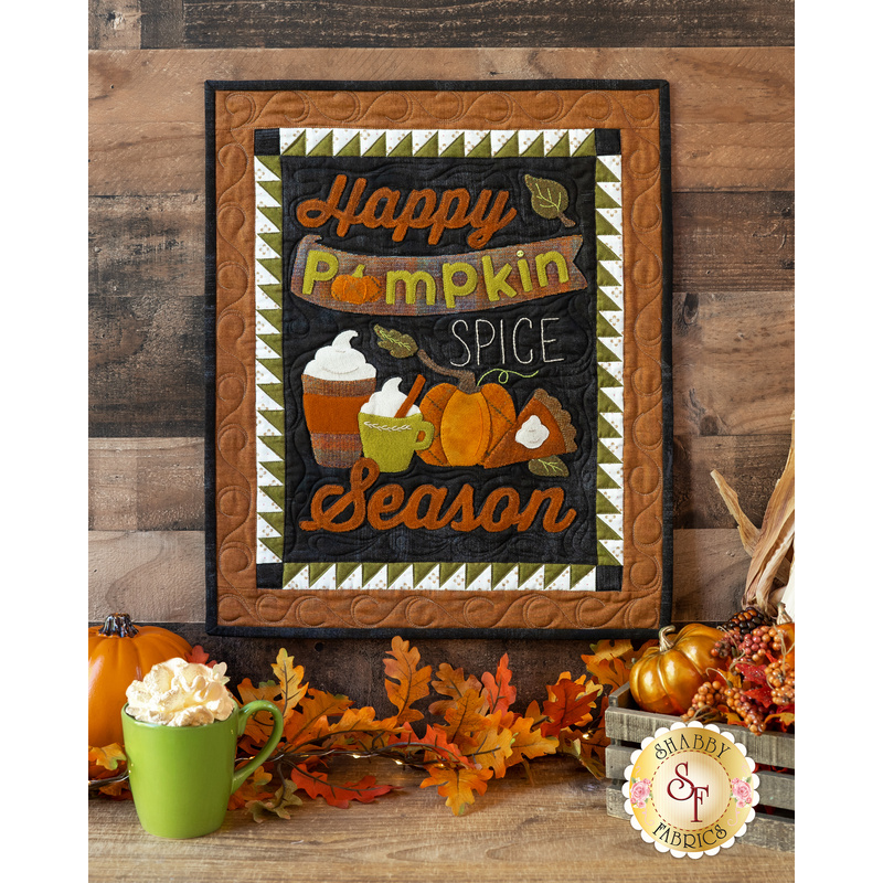 An autumn themed wool wall hanging with applique words, pumpkins, and coffee cups hung on a wooden wall