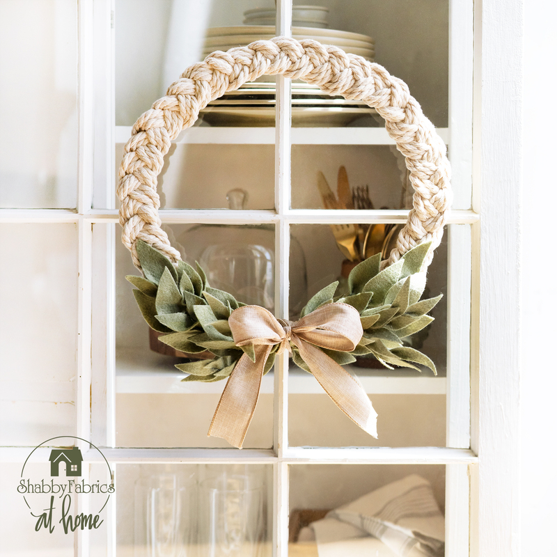 Beige wreath made with a braided design and green leaves made of fabric accented with a beige ribbon.