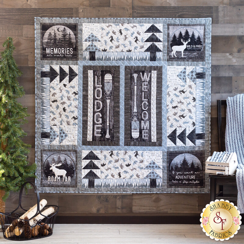 A gray, monochrome, wilderness and camping-themed, square quilt hanging on a dark paneled wall with a pine tree and basket of firewood on the left and a chair with a blanket and books on the right.