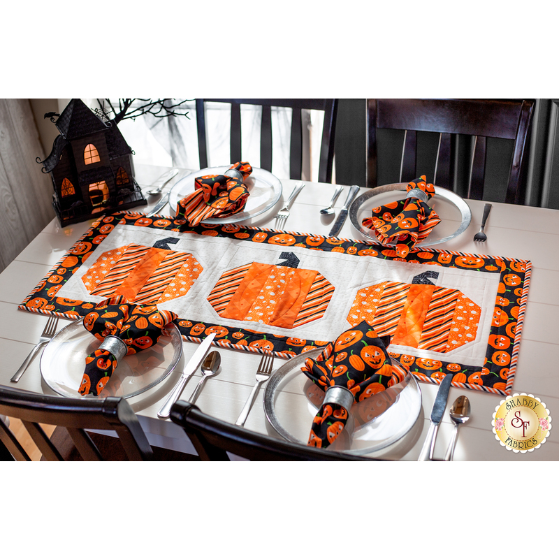 Table runner featuring 3 pumpkins made of strips of orange and black halloween themed fabrics on a table set with plates and matching napkins.