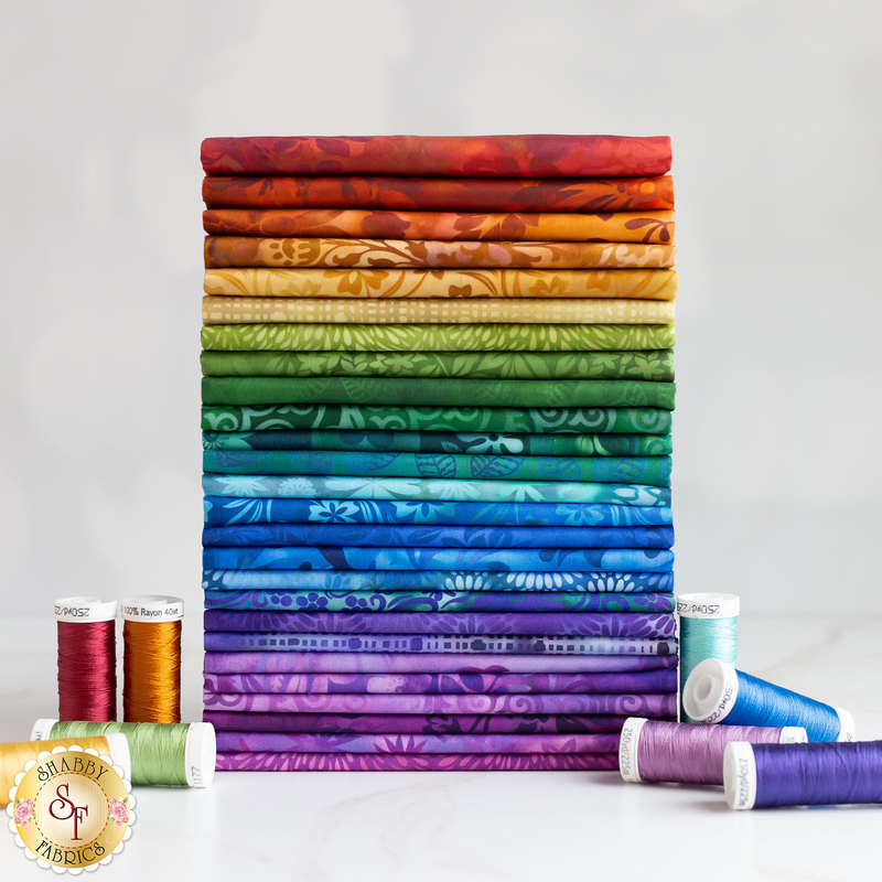 Photograph of a stack of fabrics in rainbow order against a light grey backdrop with matching spools of thread nearby