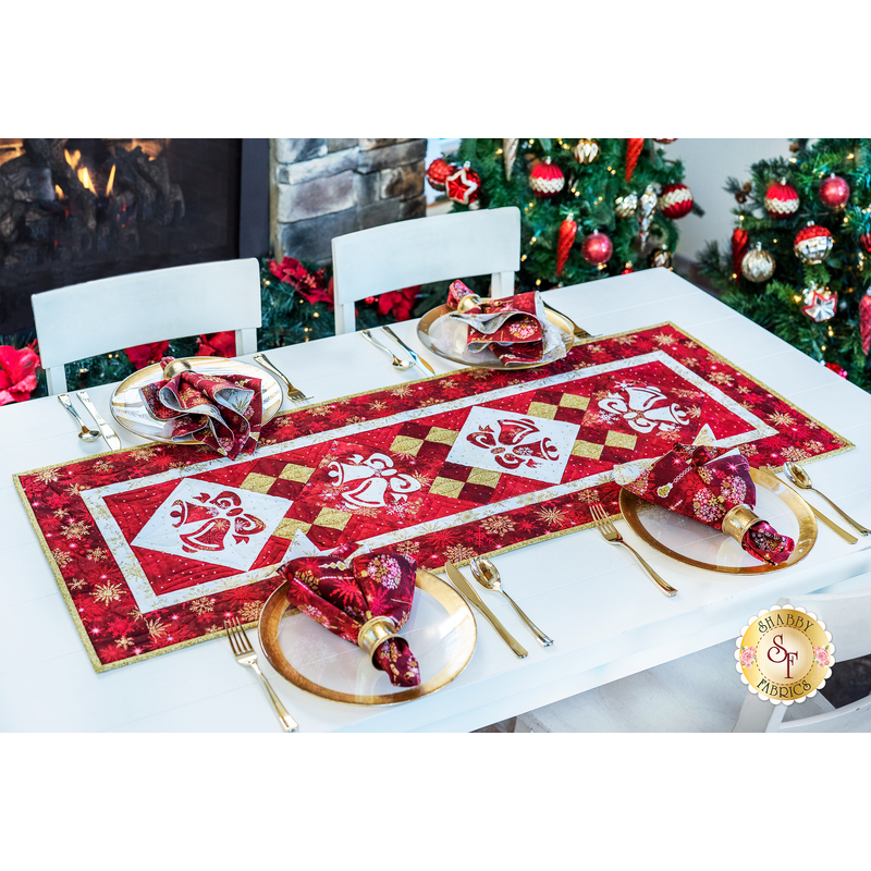 Christmas themed table runner featuring bell designs made of red and cream snowflake fabrics on table with matching napkin place settings.