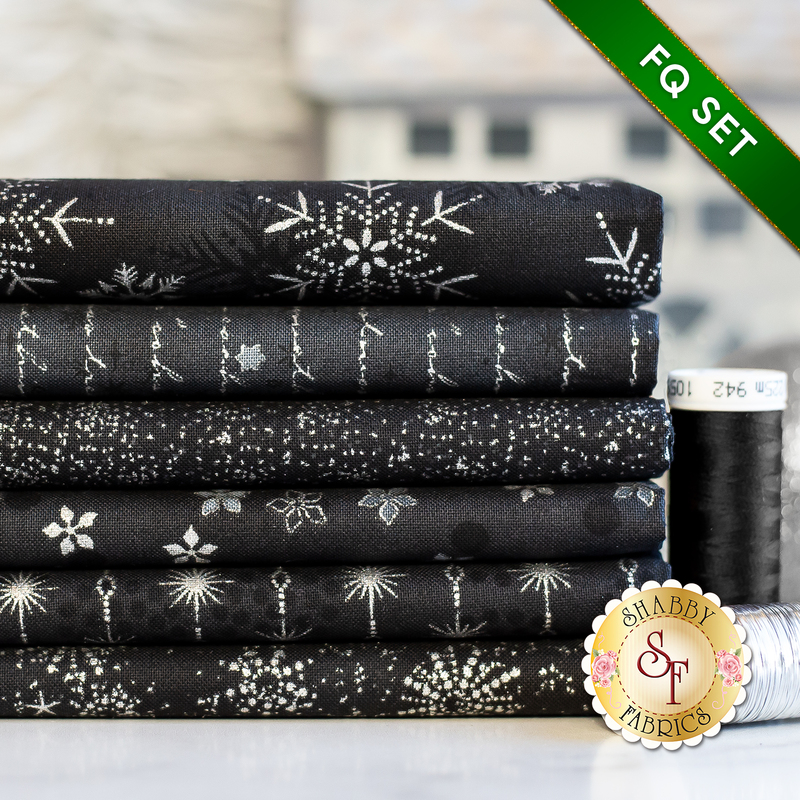Stack of 6 folded black fabrics with metallic silver snowflakes, script, and stars/burst patterns with a spool of black thread next to it on a white countertop with a pale room in the background and a green banner in one corner that reads: 