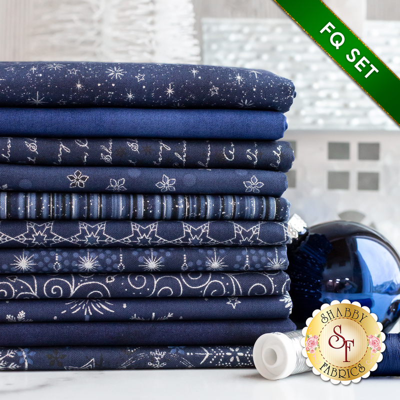 Stack of 11 folded navy blue fabrics with metallic silver snowflakes, script, swirls, and stars/burst patterns with a spool of silver thread and a large blue bauble next to it on a white countertop with a pale room in the background and a green banner in one corner that reads: 