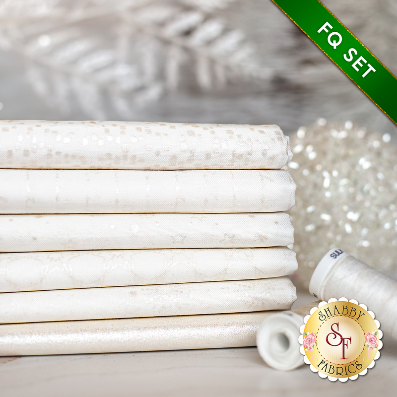 Stack of 6 folded white fabrics with pearlescent snowflakes, script, and stars/burst patterns with spools of white thread next to it with a white sparkly bauble on a white countertop with a pale decor in the background and a green banner in one corner that reads: 