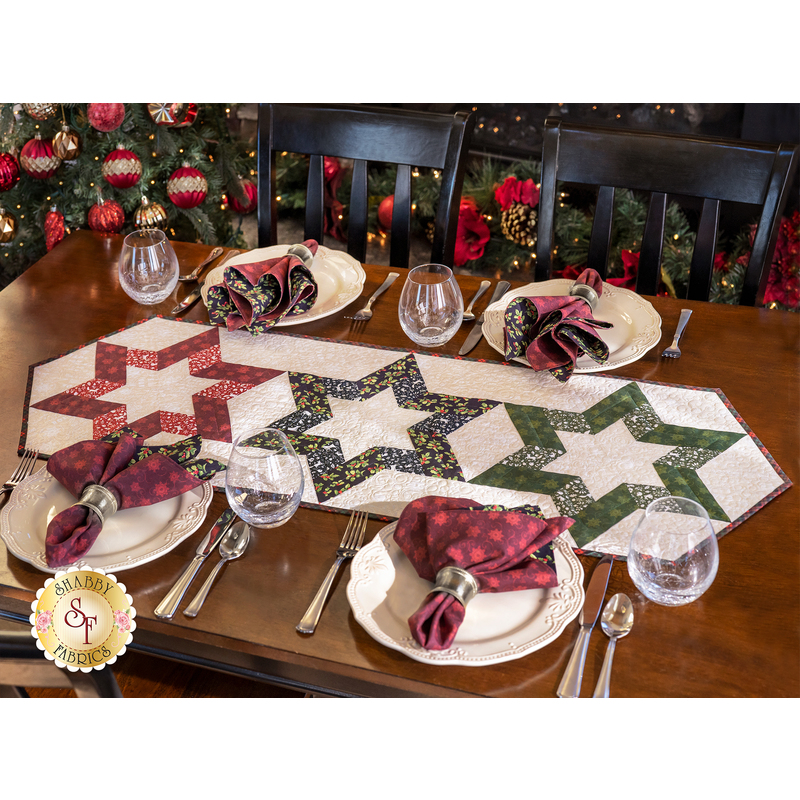 Table runner with 3 hollow star design made of red, black, and green winter theme printed fabric on cream.