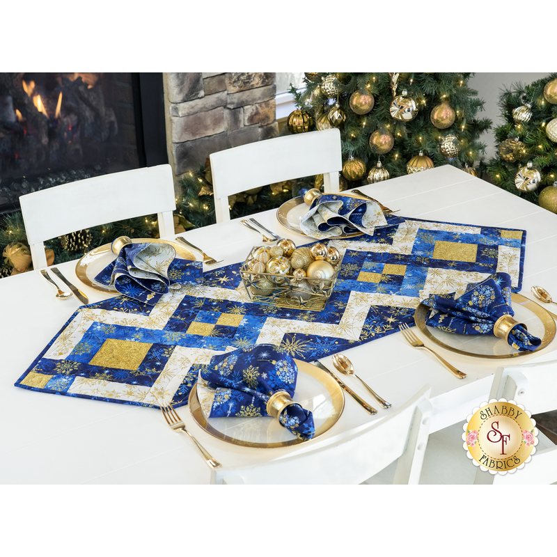 white table and chairs with christmas trees and a fireplace in the background, featuring a blue and gold table runner with gold place settings and matching napkins with a centerpiece of gold ornaments in a bowl