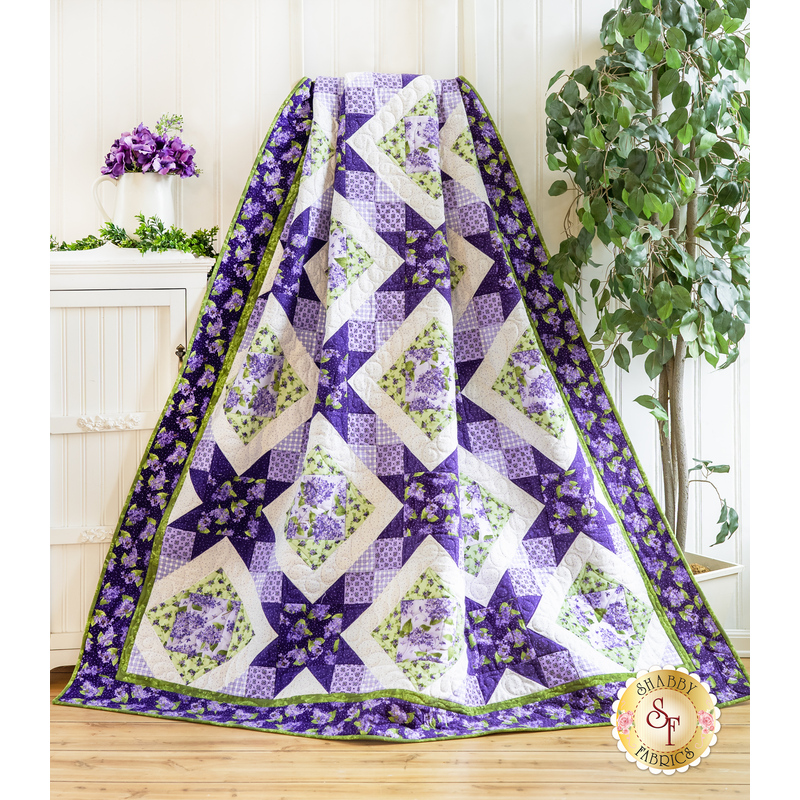 A quilt with sawtooth star design with geometric patchwork in Purple, white, and green floral fabrics draped over furniture.