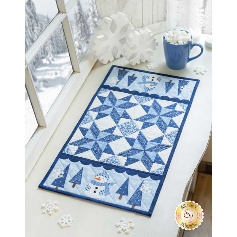 Blue and white winter themed table runner atop a white counter with a wintry scene outside an adjacent window and snowflake decorations with a mug of hot cocoa