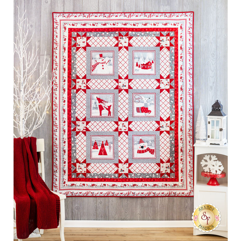 White, red and gray quilt hanging on a gray paneled wall with a small white chair with a red blanket draped over it and a small white shelf with winter decor in the foreground