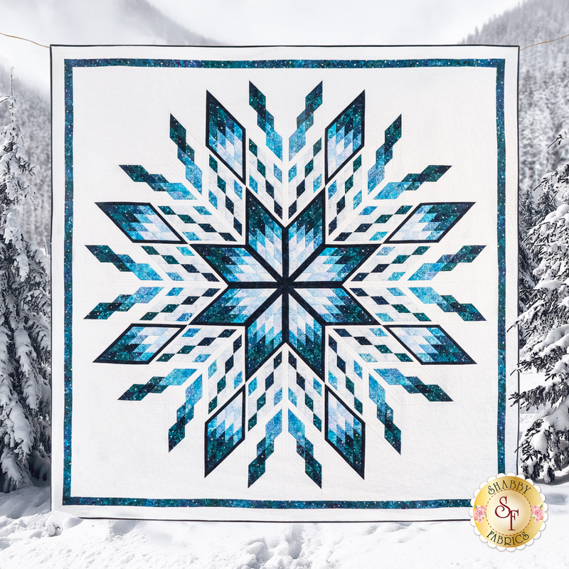 completed ice castles quilt featuring a blue starburst or snowflake-like geometric pattern on white, with snow covered evergreens and mountains in the background