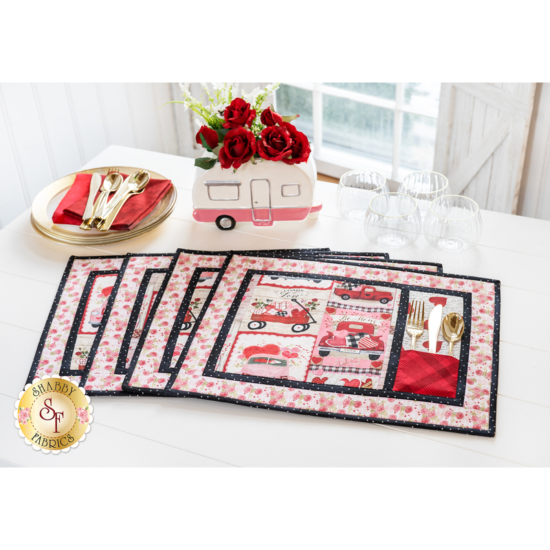 Set of 4 placemats fanned out on a white table in front of a window with a small camper flower pot decoration with red flowers and a stack of plates with red napkins and silverware