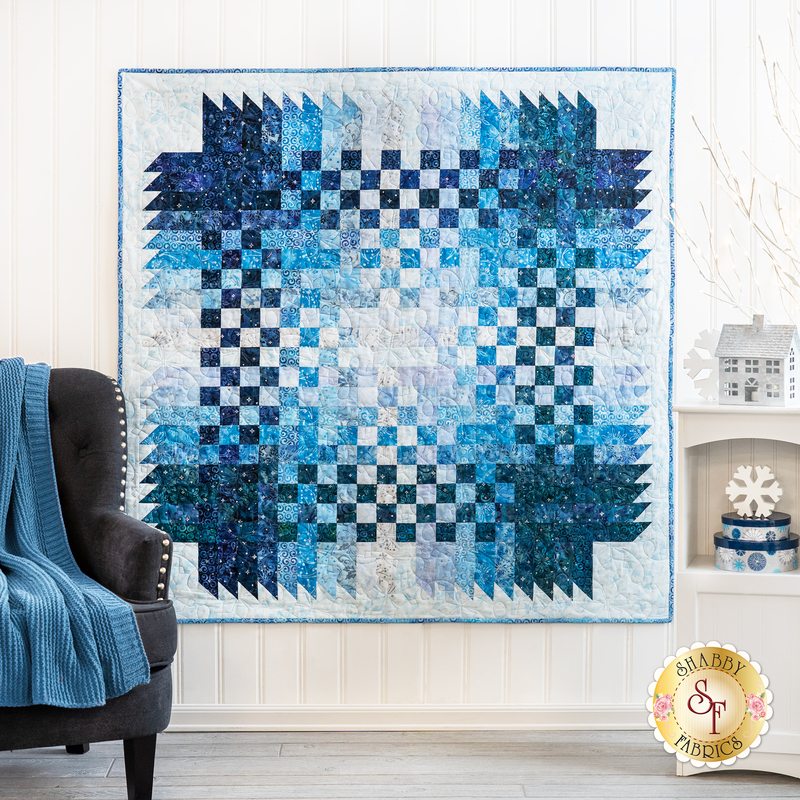 Image of finished Over and Down Under quilt in a gradient of blues and white in a woven pattern on a white wall with a chair and blue blanket with a small shelf and winter decorations in the foreground