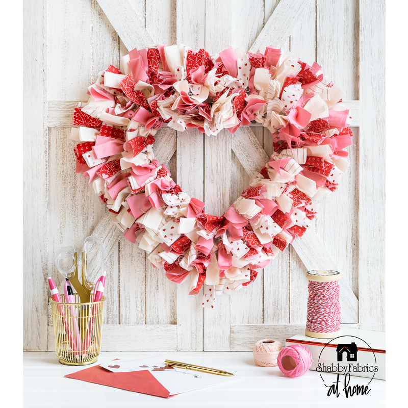 Heart shaped scrappy wreath made up of pink, red, and white fabric strips hanging on a white wooden wall with cross beams and crafting supplies and a pen holder on the white countertop in the foreground.