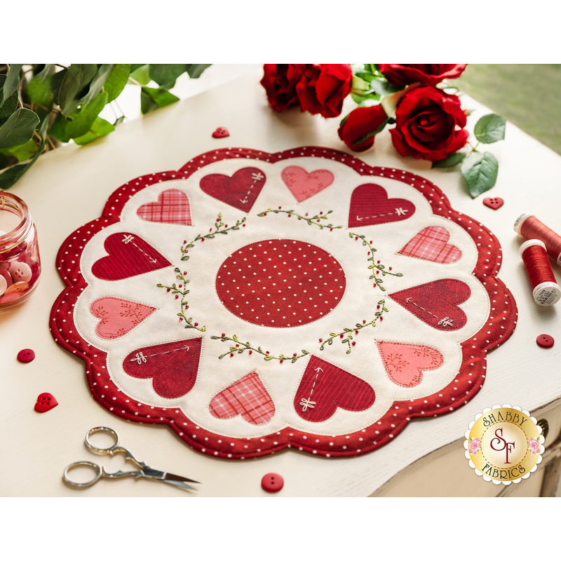 Round white table topper with scalloped edges and red and pink hearts in a circle around it, on a white countertop with red buttons, red thread, red roses, and scissors with a plant in the background