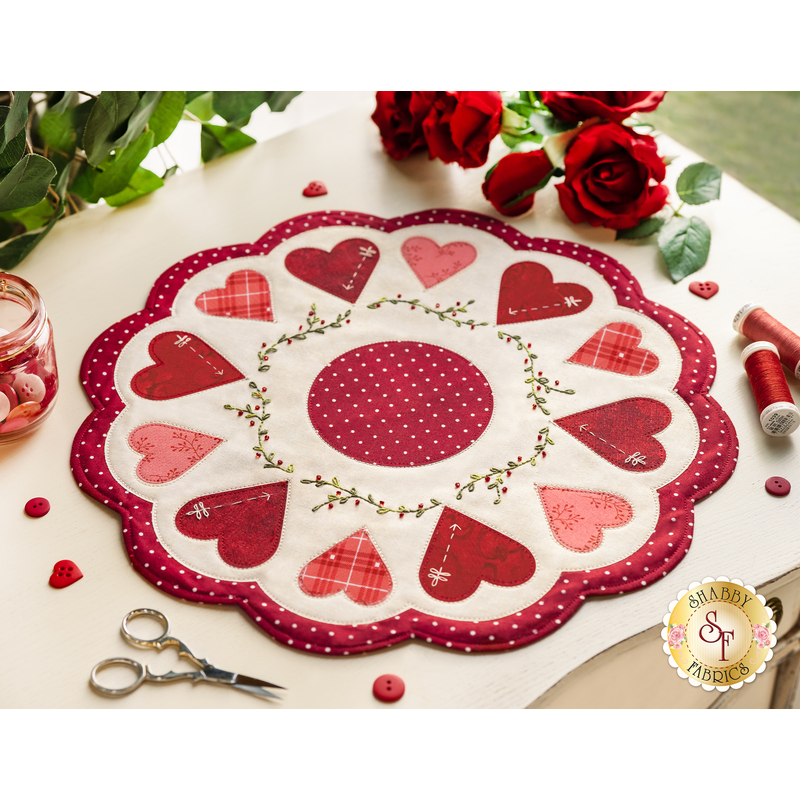 Round white table topper with scalloped edges and red and pink hearts in a circle around it, on a white countertop with red buttons, red thread, red roses, and scissors with a plant in the background
