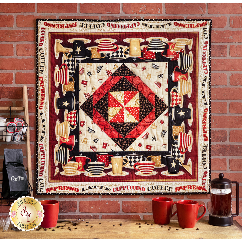Geometric small quilt featuring Coffee Always fabrics in red, black and cream, hanging on a brick wall over a coffee bar with coffee beans scattered, a french press, a rack of coffee additives, and three red mugs