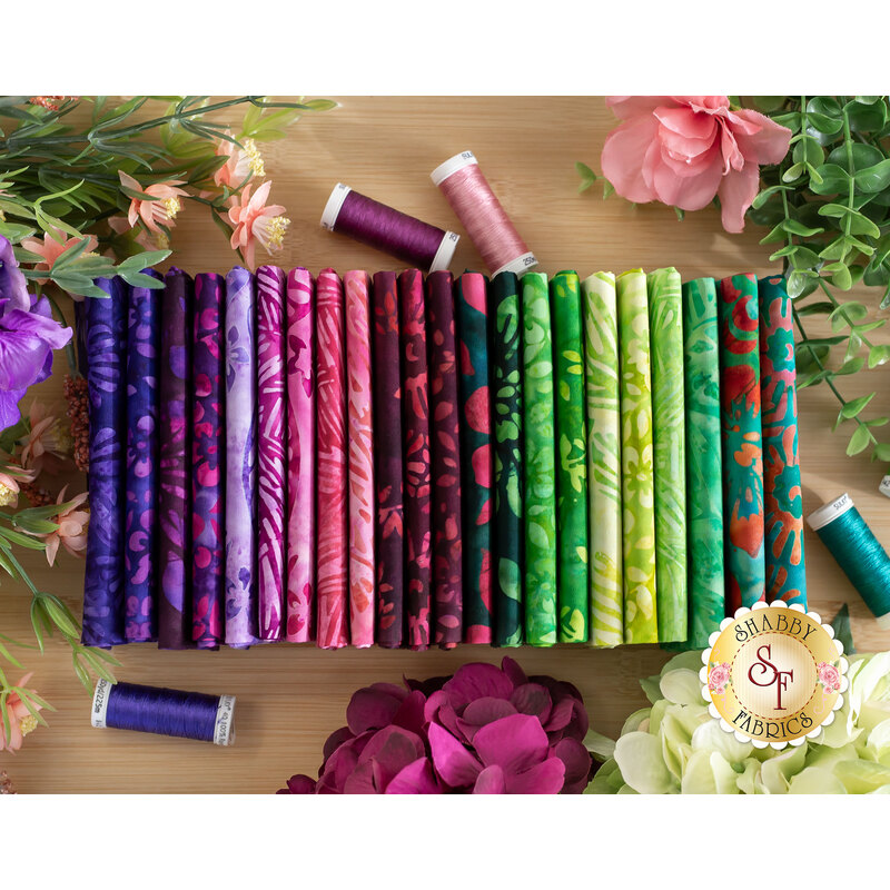 floral batik fabrics in purple, pink, and green shades, on a table surrounded by flowers and thread