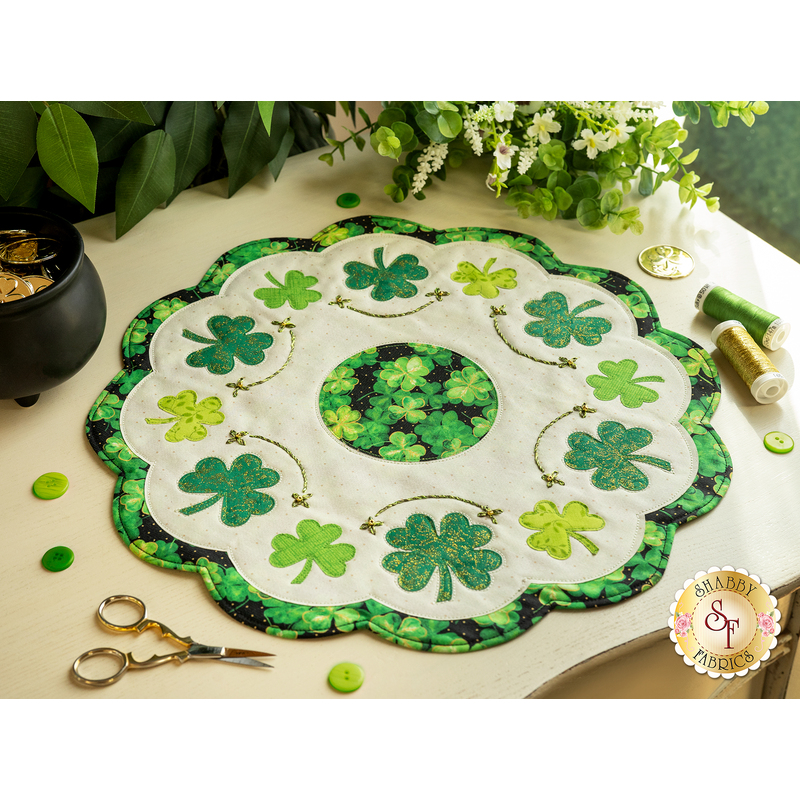 Round table topper with green shamrocks and white fabrics with hand embroidery details on a white countertop with green buttons, gold scissors, and matching threads with house plants in the background with a small black cauldron with gold inside.