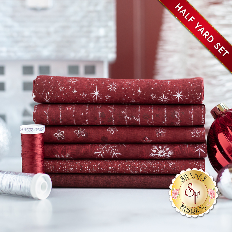 Stack of 6 folded dark red fabrics with metallic silver snowflakes, script, and stars/burst patterns with spools of red and silver thread and a large red bauble next to it on a white countertop with a pale room in the background and a red banner in one corner that reads: 