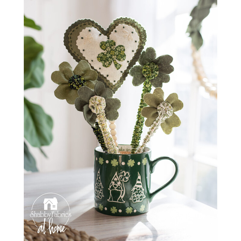 A green mug decorated with white outlines of gnomes with shamrock accents filled with decorative shamrocks and a large stuffed heart on a beaded dowel with embroidery and applique embellishments