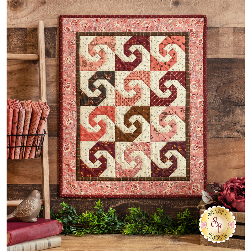 Pink, brown and cream mini quilt with blocks featuring a swirling pattern with different fabrics in each one hanging on a brown paneled wall above a brown countertop with fresh garland, books, red flowers, and a small basket of fabrics from the Chocolate Covered Cherries collection on either side.