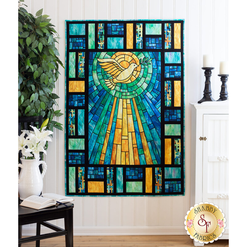 Image of a pieced quilt made of blue, yellow and aqua stained-glass looking fabric with a yellow dove and sunrays in the middle. The quilt is hanging on a wall, framed on the right by a cabinet with candles, and on the left by a tree and table with a vase on it