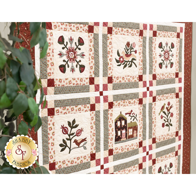 Photo of a square quilt featuring a house and floral motifs in each block hanging on a gray wall with a green houseplant in the foreground
