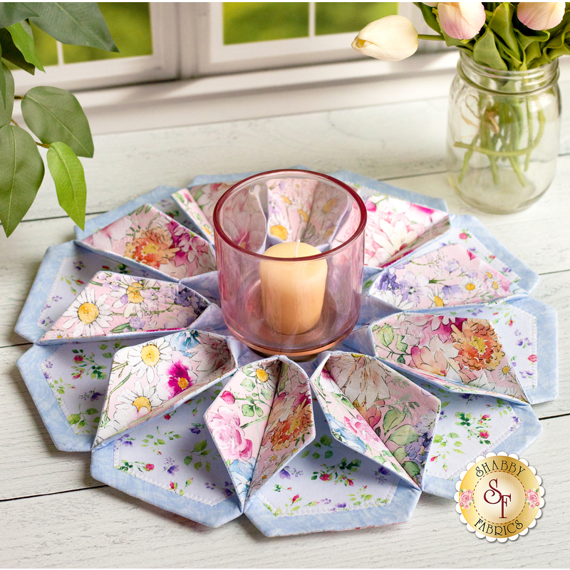 Flower shaped table topper with central opening containing a candle. Topper is made of white, soft pink and blue fabrics with multicolor florals.