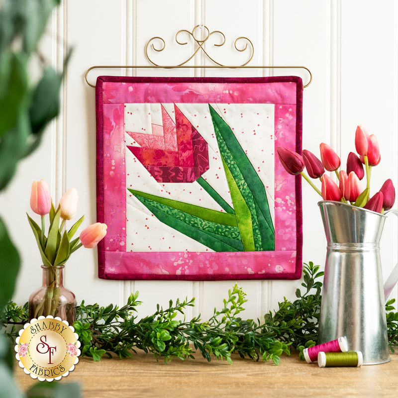 Finished quilt block featuring a pink tulip with green leaves bordered by pink mottled fabrics hanging from a craft holder decorated with curls on a white paneled wall with tulips in a metal pitcher to the left, coordinating spools of thread on the wooden countertop below, with greenery and tulips in a vase.
