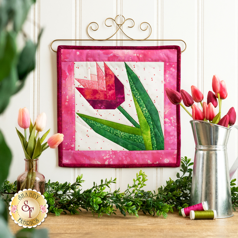 Finished quilt block featuring a pink tulip with green leaves bordered by pink mottled fabrics hanging from a craft holder decorated with curls on a white paneled wall with tulips in a metal pitcher to the left, coordinating spools of thread on the wooden countertop below, with greenery and tulips in a vase.