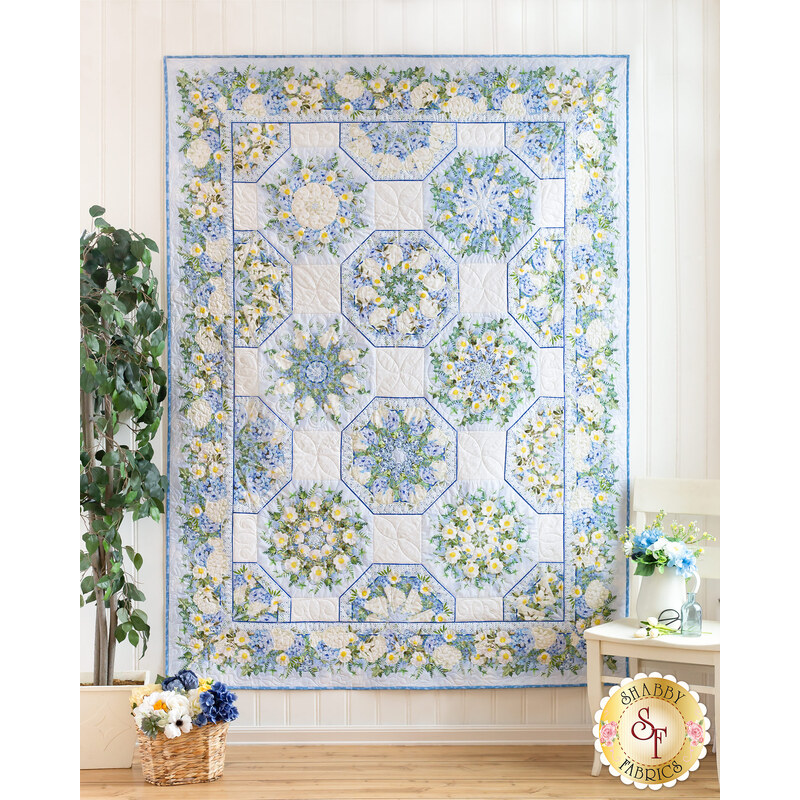 Photo of a blue, cream, and green floral quilt with octagonal blocks hanging on a white paneled wall with a houseplant and small white chair with blue flower decorations