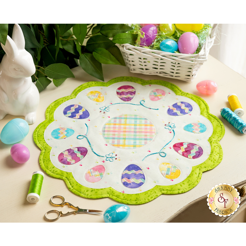 A round scalloped table topper with decorated easter eggs on a white background with a bright green border and hand embroidered details on a cream countertop with scattered notions and easter eggs all over with a ceramic rabbit and green houseplant with an easter basket in the top right corner.