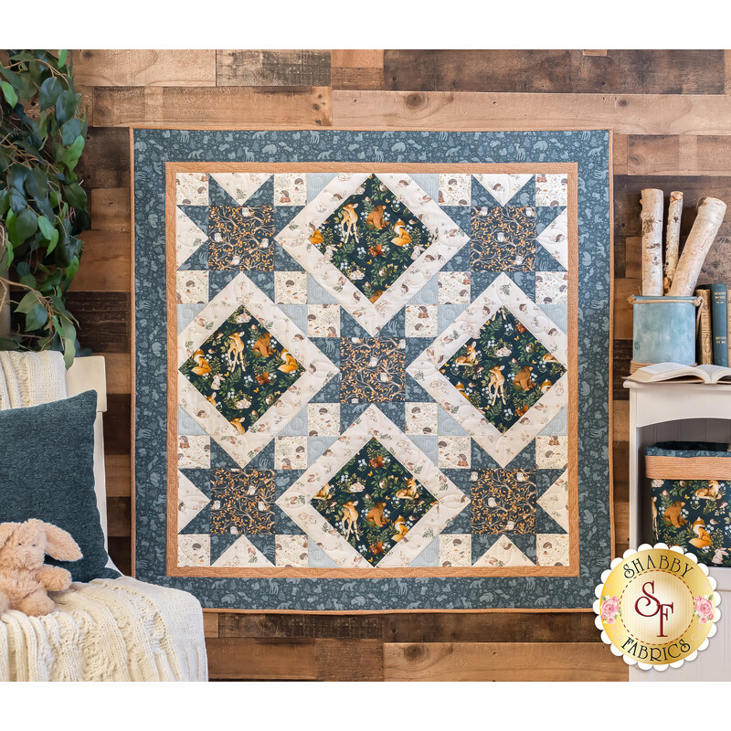 a small blue and white quilt with sawtooth star shapes and square diamonds made with the Little Forest fabric collection hanging on a brown paneled wall with a small shelf, blanket covered chair, and house plant in the background