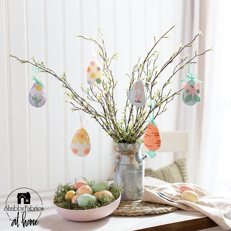 a vintage style tin milk jug holding decorative branches with 6 Easter Egg ornaments in various designs on a table with a basket full of easter eggs in the foreground in front of a white paneled wall