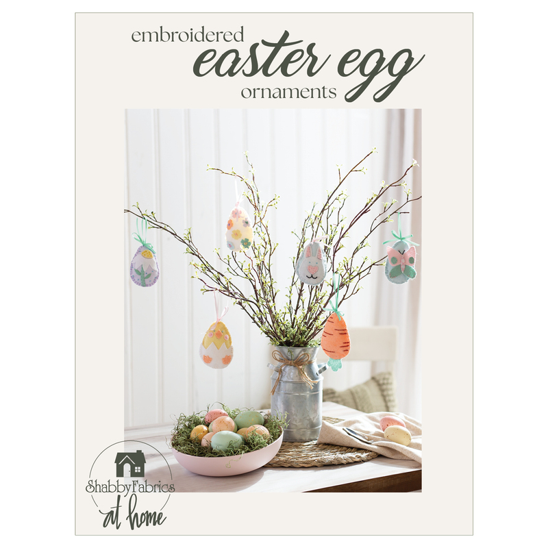 Six felt Easter egg ornaments hanging from an arrangement of spring buds on a table next to a bowl of colored eggs