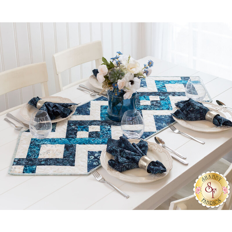 Blue and cream table runner with a criss crossing diamond pattern from end to end with pointed ends on a white wood table with four place settings with matching blue napkins and a white paneled wall with a bright window in the background